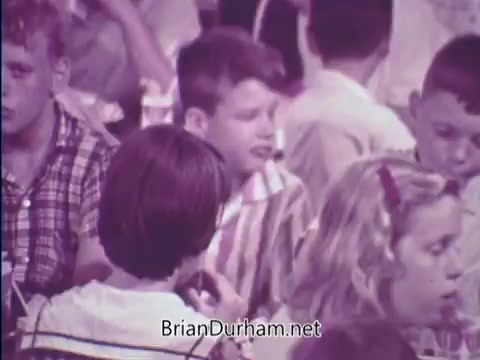 The still depicts a scene from the 1967 PSA for the USDA School Lunch Program, featuring a diverse group of children in a cafeteria setting. They are engaged in conversation and eating, with some wearing plaid and striped shirts. The image conveys a sense of community and emphasizes the importance of school lunches during that era.