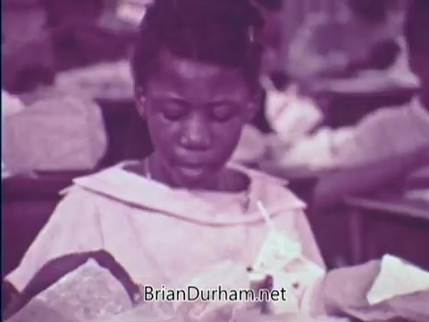 The still from the 'USDA School Lunch Program PSA - 1967' shows a young child focused on a school lunch, seated at a desk. The image is tinted in shades of purple, typical of the era. The child appears to be inspecting their meal, which includes a carton of milk, and is dressed in a school uniform or apron. The background suggests a classroom setting filled with other students and lunch items, emphasizing the context of school nourishment programs.