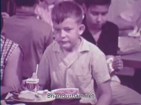 The still from the 1967 PSA on the USDA School Lunch Program shows a young boy seated at a cafeteria table, looking contemplative while holding a tray with a balanced meal. He has short hair and is wearing a light-colored, collared shirt. In the background, other children are present, engaged in eating and conversation, highlighting a typical school lunch environment of that era. The scene emphasizes the importance of nutrition for school children.