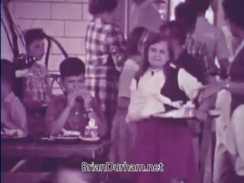 The still from the 1967 USDA School Lunch Program PSA depicts a school cafeteria scene. Children are engaging in lunchtime activities; one girl is smiling while holding a tray of food, while a boy nearby appears to be eating. The environment features a variety of students, highlighting the social aspect of school lunches. The image reflects the educational and nutritional aims of the USDA program during that era.