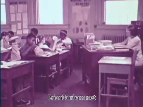 The still from the USDA School Lunch Program PSA (1967) depicts a classroom setting with children engaged in learning. There are several students sitting at wooden desks, focusing on their tasks, while a teacher instructs from a central desk. The environment appears bright and educational, with charts and educational materials visible on the walls, emphasizing the theme of school nutrition and education.