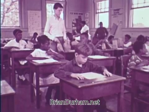 The still shows a classroom scene, likely from the 1967 USDA School Lunch Program PSA. Several children are seated at wooden desks, engaged in their work. A teacher, dressed in a white shirt, is interacting with a student in the foreground, while others appear focused on their tasks. The classroom has large windows that allow natural light, and educational materials are visible on the walls. The atmosphere conveys a standard school setting, emphasizing education and the involvement of educators.