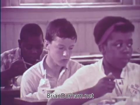 The still from the 'USDA School Lunch Program PSA' (1967) features two children eating lunch in a school setting. The focus is on a boy with short hair, appearing thoughtful, and another child, a girl, who is actively eating. The scene conveys the program's emphasis on providing nutritious meals to students and highlights diversity among the children. The overall tone is one of camaraderie in a nurturing environment.