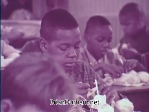 The still from the 1967 USDA School Lunch Program PSA depicts a group of schoolchildren seated at tables, focused on their lunches. One boy, prominently featured in the foreground, appears engaged in his meal, showcasing the initiative's emphasis on providing nutritious food to students. The overall tone reflects the importance of school lunches in promoting children's health and well-being.