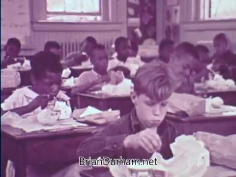 The still depicts a classroom scene from the 1967 USDA School Lunch Program PSA. In the image, several children are seated at desks, eating their lunches, which appear to consist of wrapped sandwiches. The focus is on a young boy in the foreground, who is intently eating, while other children engage in their meals as well. The atmosphere suggests a typical school setting, highlighting the program's intention to provide nutritious meals to students. The color palette gives it a vintage look, reflective of the time period.