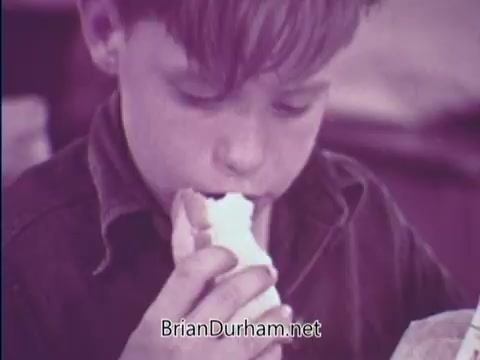 The still features a young boy, intently eating a sandwich. The image has a vintage quality, characterized by a muted color palette, likely emphasizing the educational theme of the USDA School Lunch Program in the context of the late 1960s. The boy's focused expression suggests enjoyment and emphasizes the importance of nutritious meals provided by the program.