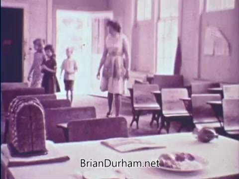 The still from the 16mm PSA for the USDA School Lunch Program features a classroom setting with wooden desks and chairs. A woman, presumably a teacher or caregiver, interacts with three children—a girl in a dress, another girl, and a boy. The scene conveys a sense of community and education, highlighting the importance of school meals. The overall ambiance is simple and reflective of the 1960s, with soft colors and a focus on children's well-being.
