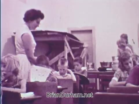 The still features a classroom scene from the 1967 USDA School Lunch Program PSA. A woman, likely a school staff member, is interacting with children at their desks. Some kids are engaged in activities, while others appear to be focused on their work. In the background, a large table with food items is visible, suggesting an emphasis on nutrition and the school lunch initiative. The overall atmosphere reflects a typical educational environment of that era.