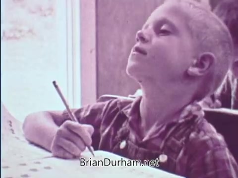 The still features a young boy, focused and engaged, seated at a desk while holding a pencil. He appears to be in a classroom setting, possibly participating in an educational activity related to the USDA School Lunch Program. The boy's expression suggests concentration and thoughtfulness as he works on his task. The overall tone of the image supports a theme of education and nutrition important at the time.