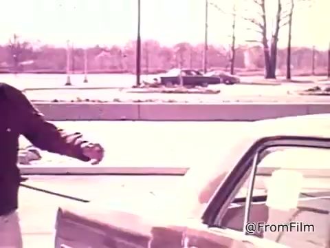 The still from 'New Federal Fuel Economy Standards 1975' shows a person standing next to a car in a parking lot, appearing to check or interact with the vehicle. The scene captures a moment emphasizing the transition to considering fuel economy in car purchases, highlighting the importance of gas mileage information, as indicated by the window sticker. The background shows a sparse parking area, suggesting a typical day during the 1970s.