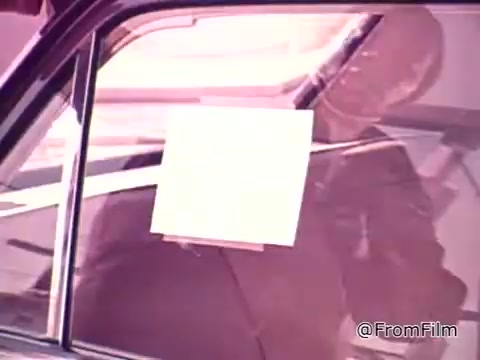 The still from the 16mm film 'New federal fuel economy standards 1975' shows a close-up of a car window with a white sticker affixed to it. The sticker likely details the vehicle's fuel economy ratings for city and highway driving. The context emphasizes the rising costs of gas and the importance individuals place on fuel efficiency when selecting a new car.