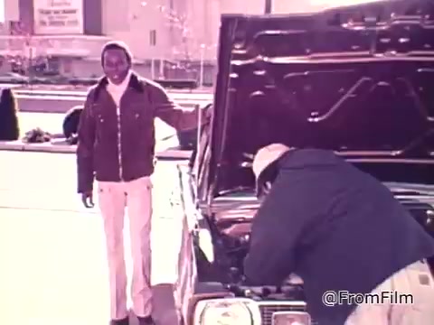 The still features two individuals near a car, with one person standing by the hood while another examines the engine. The context relates to rising fuel prices and the importance of understanding fuel efficiency, highlighted by the discussion of gas mileage stickers on new cars, emphasizing conservation and awareness in vehicle purchases.