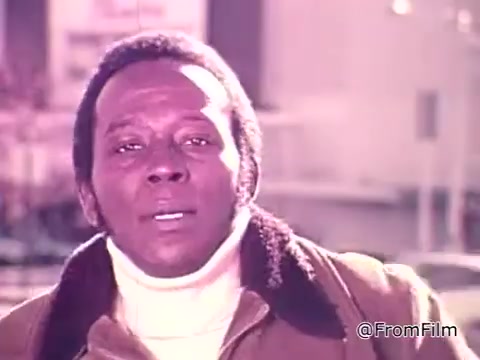 The still features a man speaking directly to the camera, emphasizing the importance of fuel efficiency and gas conservation in the context of rising fuel prices. He notes how he considers gas mileage when purchasing a new vehicle, specifically referencing the window stickers that provide information on city and highway fuel economy. The setting suggests a focus on consumer awareness and environmental consciousness reflective of the 1975 era and the new federal fuel economy standards.