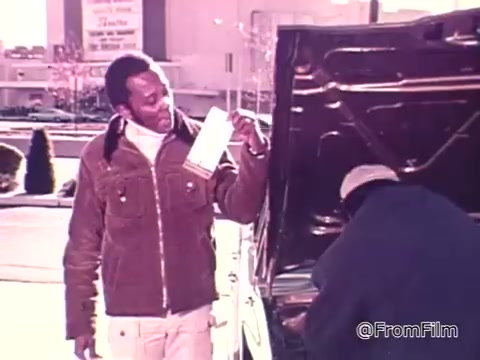 The still shows a man holding up a window sticker with gas mileage information, reflecting on rising fuel costs. He stands near a car with its hood open, while another person works under the hood. The setting suggests a focus on car efficiency and the importance of gas conservation amid the 1975 fuel economy standards.