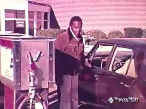 The still depicts a man standing next to a gas pump, engaging with his vehicle. He seems to be focused on the cost of fuel and the importance of gas mileage. The context highlights the rising gas prices in the mid-1970s, prompting consumers to consider fuel efficiency when purchasing vehicles. The mention of the gas mileage sticker reflects a new awareness about conserving fuel during that era.