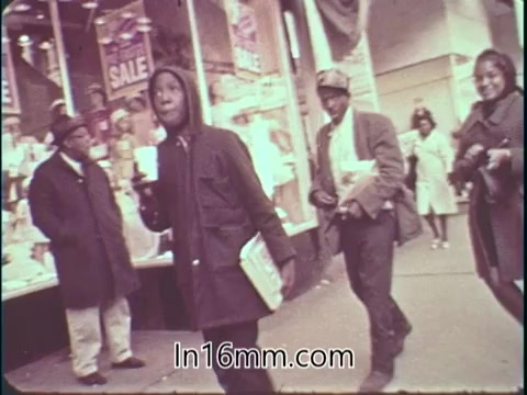 The still from the 'College Fund PSA - 1960's' features a vibrant street scene showcasing a group of individuals. In the foreground, a young man in a hooded jacket holds a cup while walking, exuding energy. Two others, a man in a hat and a woman, follow behind, highlighting a sense of community. Storefronts in the background display sale signs, adding to the bustling atmosphere of the era. The overall tone reflects the optimistic spirit of youth and the importance of education.
