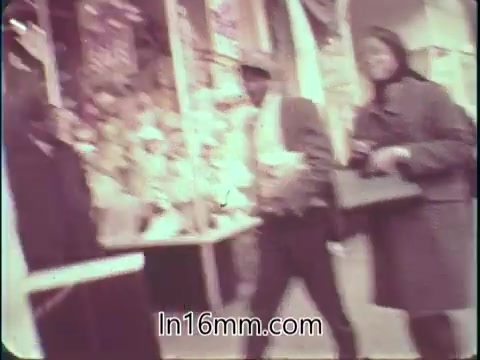 The still from 'College Fund PSA - 1960's' shows a street scene with pedestrians walking by. The image features a man in a hat and a woman in a coat, both engaged in their activities. Background elements include colorful advertisements or banners, indicative of a busy urban environment during the 1960s. The overall tone is candid and vibrant, reflecting the era's ambiance.