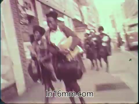 The still from the 'College Fund PSA - 1960s' features a group of students, likely on their way to or from school. They are dressed in 1960s attire, including coats and carrying books and bags, conveying a sense of urgency. The urban setting suggests a school environment, highlighting the importance of education and the college fund message. The image has a warm color palette typical of the era.