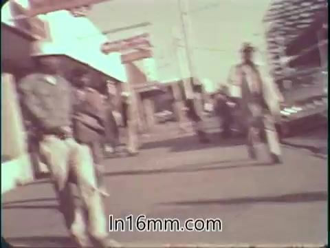 The still depicts a street scene from the 1960s, showing pedestrians casually walking along a sidewalk. The environment appears informal, with storefronts visible in the background. The colors are muted, likely due to the film technology of the time, contributing to its nostalgic feel. The subjects in the image are dressed in period-appropriate clothing, reflecting the era's fashion trends.