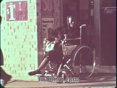 The still from the 'College Fund PSA - 1960's' shows a person sitting in a wheelchair outside a storefront. The scene features a brick-patterned wall and a visible window, with various signs promoting low prices. The overall mood appears reflective, emphasizing themes of accessibility and support for education.