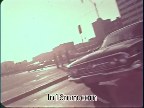 The still depicts a 16mm film scene from a 1960s college fund PSA. It features a vintage car parked in the foreground, with a cityscape in the background, including buildings and cranes. The lighting appears warm, suggesting a sunny day, and the scene conveys a mid-century urban environment. The composition emphasizes the car and the city context, likely promoting higher education and its opportunities.