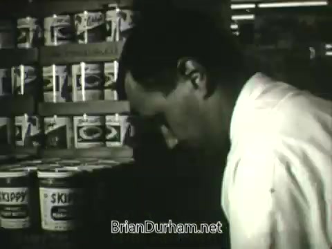 The 16mm still from the 'Hire the Handicapped PSA' features a man in a white shirt engaged in work, presumably in a store setting. The background shows shelves stocked with various cans, emphasizing a retail environment. The surrounding context suggests a strong message about inclusivity and the capabilities of veterans and individuals with disabilities, encouraging viewers to recognize their potential and contributions to society.
