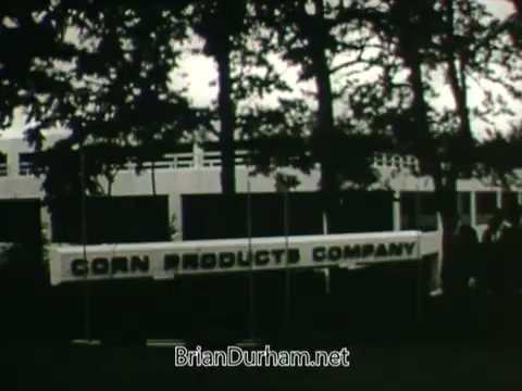 The still from the 'Hire the Handicapped' PSA features a prominent banner with the text 'CORN PRODUCTS COMPANY,' set against a backdrop of trees. The image conveys a message of support for veterans and individuals with disabilities, emphasizing their eagerness to learn and contribute to the workforce. The PSA encourages viewers to consider how they can help these individuals by providing opportunities, promoting the idea that everyone has abilities worth recognizing.