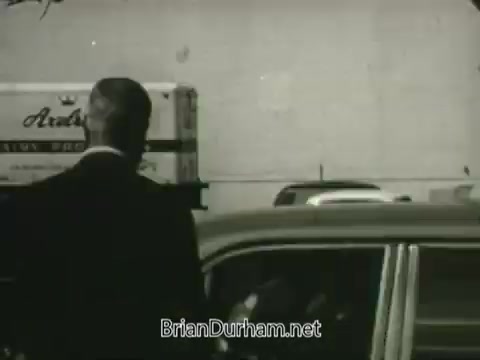 The still from the 'Hire the Handicapped' PSA features a man in a suit, seen from behind, standing next to a vintage car. Behind him, there is a delivery truck with the words 'Aradale' visible. The setting appears to be urban, likely in the mid-20th century, reflecting a call to action to support veterans and differently-abled individuals in the workforce. The message emphasizes recognizing abilities rather than disabilities, promoting inclusivity and opportunities for these individuals.