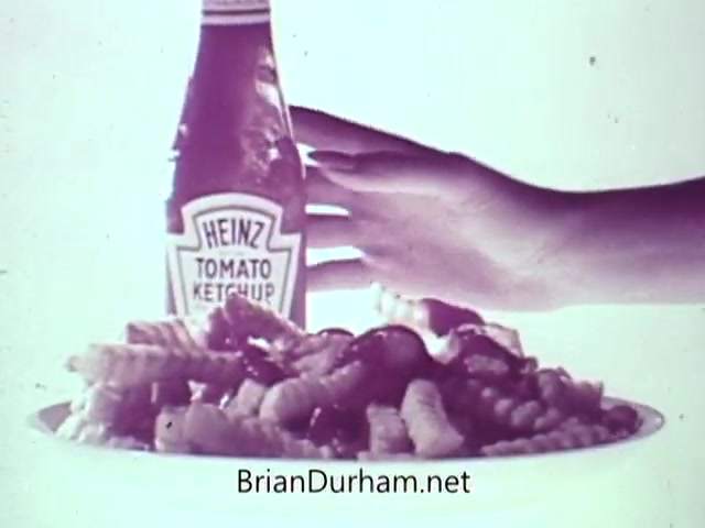The still depicts a bottle of Heinz Tomato Ketchup prominently positioned next to a plate of pasta. A hand reaches toward the ketchup, emphasizing its use as a condiment. The visual conveys the brand's iconic status in the 1960s, highlighting its association with food enjoyment, particularly with pasta dishes. The aesthetic suggests a playful, nostalgic appeal, likely aimed at drawing viewers' attention to the product's unique qualities, such as its slow pour.