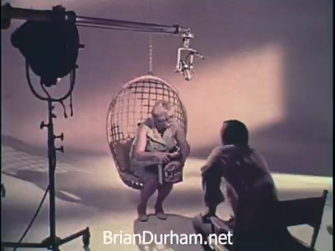 The still features a woman sitting in a hanging wicker chair, engaged in a conversation with a crew member off-screen. She's holding a box of Oxydol laundry detergent, suggesting a relaxed yet promotional atmosphere. The set is minimally decorated with studio lighting equipment, indicative of a 1960s commercial shoot. The overall aesthetic reflects the era's advertising style, emphasizing comfort and household care.