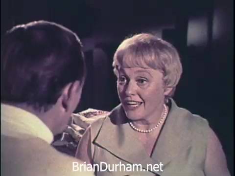 The still features a conversation between two characters, likely in a domestic setting, reflecting the style of 1960s television commercials. The woman, with a friendly demeanor, is wearing a light-colored outfit and pearl necklace, suggesting a typical housewife image of the era. The focus is on the dialogue, likely promoting Oxydol laundry detergent, emphasizing its effectiveness and appeal to homemakers. The setting is warm and welcoming, indicative of the marketing style of that time.