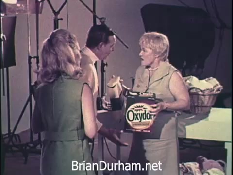 The still features a vintage Oxydol laundry detergent commercial from the 1960s. Three individuals are engaged in a discussion, with one woman holding a box of Oxydol while another holds a bottle. The setting appears to be a studio, indicated by lighting equipment and a laundry basket in the background. The scene reflects the typical advertising style of the era, emphasizing the product's benefits in a conversational context.