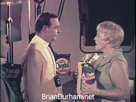 The still features a commercial from the 1960s for Oxydol laundry detergent. It shows two middle-aged individuals, a man and a woman, engaged in conversation. The man holds a box of Oxydol, while the woman, dressed in a light-colored outfit, appears to be presenting or promoting the product. A third individual is partially visible on the left. The setting suggests a studio or promotional context, typical of vintage advertisements.