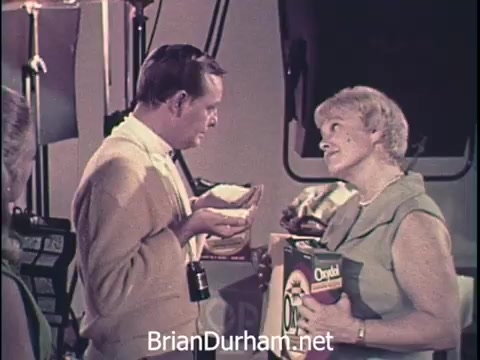 The still features a scene from a 1960s Oxydol laundry detergent commercial. It depicts a man, dressed in a sweater, holding a bowl while engaging in an enthusiastic conversation with a woman, who is holding a box of Oxydol. The setting appears to be a studio with lights in the background, suggesting a promotional shoot. The tone is friendly and persuasive, typical of advertisements from that era.