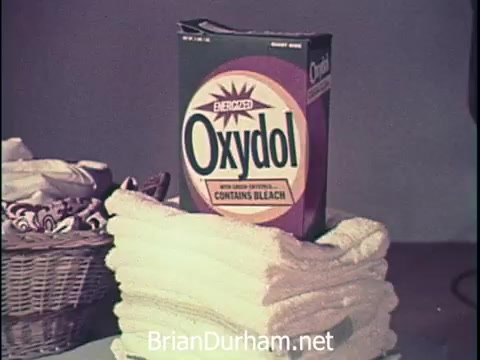 The still features a box of Oxydol laundry detergent prominently displayed on top of neatly stacked white towels. A wicker basket with additional towels is visible in the background, suggesting a clean and organized laundry setting. The packaging emphasizes that the product contains bleach, highlighting its effectiveness. The overall presentation evokes a sense of cleanliness and household efficiency typical of 1960s advertisements.