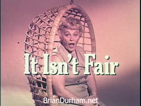 The still features a woman seated in a large woven chair, expressing surprise or frustration with the phrase 'It Isn't Fair' prominently displayed. The visual style is typical of 1960s commercials, with bright colors and engaging imagery aimed at capturing audience attention for Oxydol laundry detergent. The woman's presentation conveys a playful, relatable sentiment regarding laundry challenges.