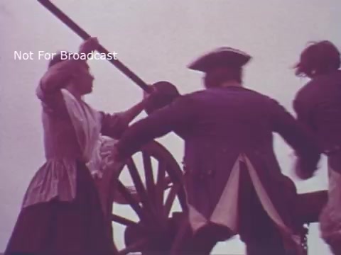 The still from the 'Molly Pitcher Bicentennial Savings Bonds PSA' depicts a historical reenactment featuring a woman, presumably Molly Pitcher, actively engaged in loading a cannon during a Revolutionary War scene. She wears period-appropriate attire, including a long dress and apron. Two men in military uniforms assist her, emphasizing teamwork and courage. The scene captures a dynamic moment, symbolizing women's contributions during the war and reinforcing themes of patriotism associated with the bicentennial celebration.