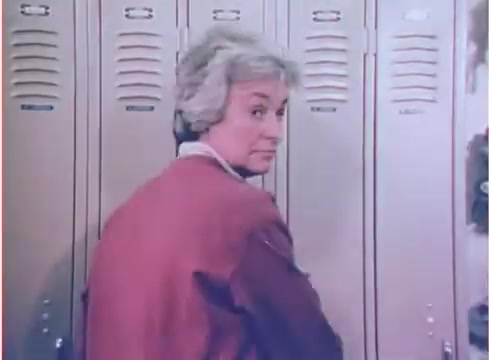 The still features a woman with short, light-colored hair, dressed in a red sweater, standing by a row of lockers. She looks back over her shoulder with a slightly inquisitive expression. The scene likely reflects a casual, everyday setting typical of the 1970s. The overall aesthetic suggests a warm color palette common in that era.