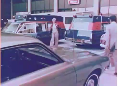 The still from a 16mm negative features a scene set in the 1970s, highlighting a parking area with several vehicles, including two vintage station wagons and a green car in the foreground. A person in light-colored clothing stands beside the parked cars, possibly discussing or demonstrating something related to oil filters. The setting appears to be a commercial or automotive environment, conveying a retro aesthetic typical of that era.