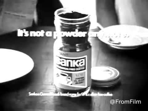 The still features a jar of Sanka decaffeinated coffee prominently displayed, with a simple, bold tagline that reads, 'It's not a powder anymore.' The image is in black and white, likely owing to the heavily red tones typical of old Eastman color film. The overall aesthetic conveys a retro vibe typical of 1960s commercials, drawing attention to Sanka's product while inviting viewers to rethink their coffee choices.