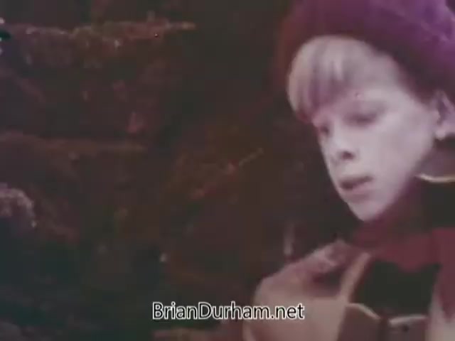 The still from 'Christmas Seal PSA - 1968' features a young boy in winter clothing, including a hat and scarf, looking contemplatively as he stands near a Christmas tree. The colors are warm and slightly faded, typical of the era's film stock. The setting suggests a holiday theme, aligning with the PSA's purpose of promoting awareness for Christmas seals.