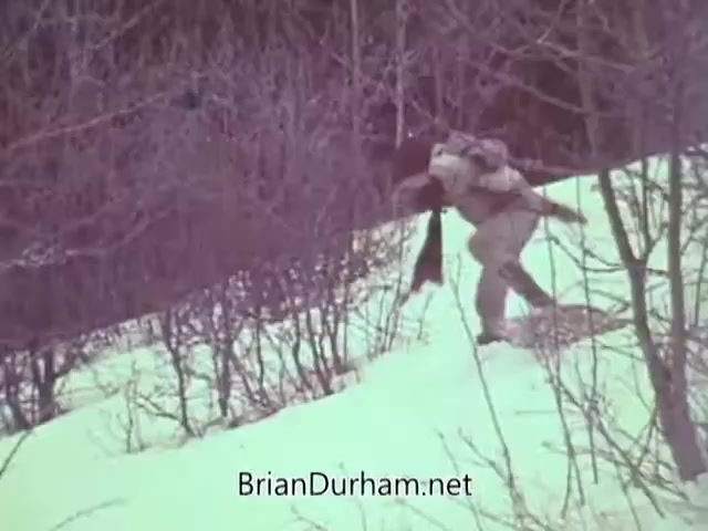 The still from 'Christmas Seal PSA - 1968' depicts an individual in winter attire navigating a snowy landscape, possibly carrying a small child or bundle on their back. Surrounding them are sparse bushes, emphasizing a barren winter scene. The image captures a moment likely intended to convey themes of care and community support associated with the Christmas Seal campaign.