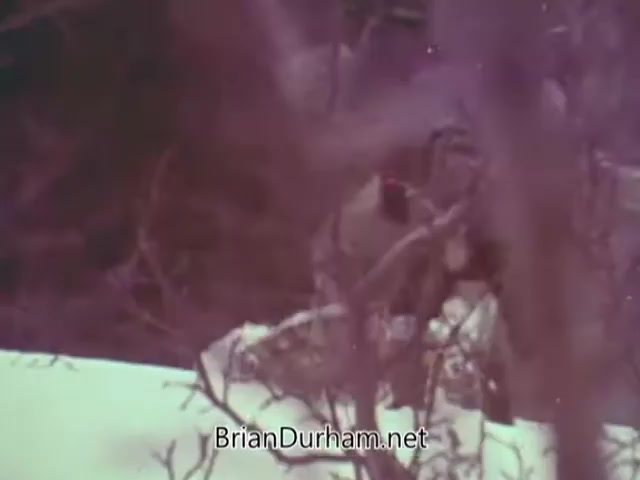 The still from the 'Christmas Seal PSA - 1968' features a snowy outdoor setting, where a figure is partially obscured by trees and branches. The atmosphere appears serene and wintery, likely conveying a message related to the holiday season and possibly the importance of supporting health initiatives associated with the Christmas Seal campaign. The color tones suggest a vintage feel typical of 16mm film.