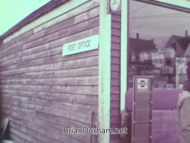 The still depicts a wooden post office building with a prominent 'POST OFFICE' sign. The exterior appears weathered, reflecting a rustic charm typical of the late 1960s. A person stands nearby, suggesting a community setting. The scene likely emphasizes the importance of mailing Christmas seals during the holiday season, as promoted by the PSA.