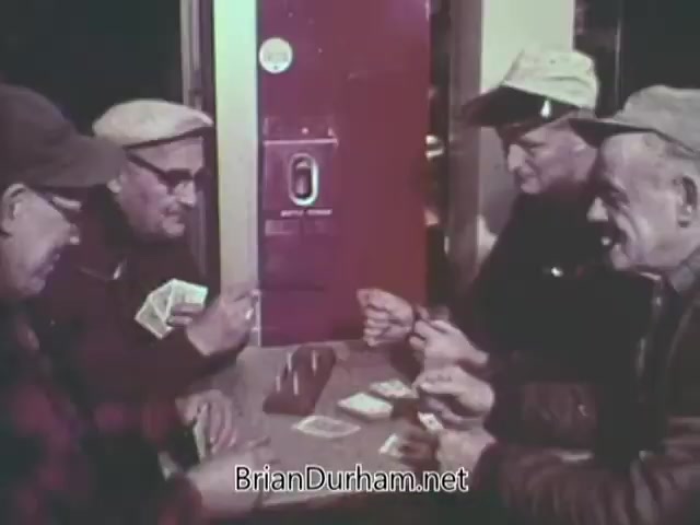 The still from the 1968 'Christmas Seal PSA' features four older men gathered around a table, engaged in a card game. They are wearing hats and appear to be enjoying each other's company, with visible camaraderie. In the background, there is a vending machine, hinting at a casual setting. The scene reflects a warm, community atmosphere, typical of holiday themes.
