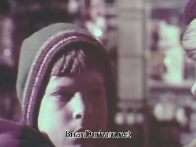The still from the 1968 Christmas Seal PSA features a young boy wearing a winter hat, with a serious expression as he appears to be engaged in conversation. The background suggests a festive atmosphere, likely among holiday decorations or goods. The focus on the boy emphasizes the emotional appeal typical of public service announcements aimed at promoting awareness and support for charitable causes during the Christmas season.