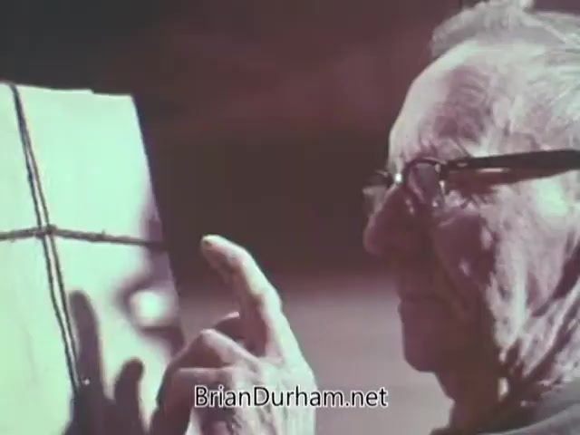 The still from the 1968 Christmas Seal PSA features an elderly man, wearing glasses, closely examining a wrapped gift or package. He points at it thoughtfully, suggesting a moment of contemplation or connection to the holiday theme. The background is softly blurred, emphasizing the man's focus on the item in front of him. This image likely reflects themes of generosity and the spirit of giving during the holiday season.