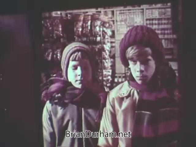 The still from the 1968 'Christmas Seal PSA' features two children standing side by side. They are dressed warmly in colorful winter clothing, including hats and scarves, suggesting a cold holiday season. The background appears to be a store display, likely filled with festive items, creating a poignant atmosphere around the theme of the PSA, which likely focuses on holiday giving and awareness for health-related causes.