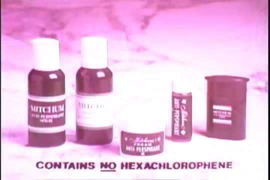 The still features a display of various Mitchum anti-perspirant products from the 1970s, arranged on a marble-like surface. Prominently placed are two bottles labeled 'Mitchum,' alongside a cream container and a stick. The text at the bottom clearly states 'CONTAINS NO HEXACHLOROPHENE.' The image has a pink tint typical of 16mm prints from that era.