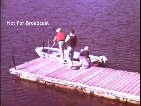 The still shows three individuals on a wooden dock near a body of water, preparing to board a small boat. Two people are wearing life jackets, emphasizing safety in boating, which aligns with the message of the PSA. The scene conveys an outdoor, recreational atmosphere with a focus on safe boating practices.
